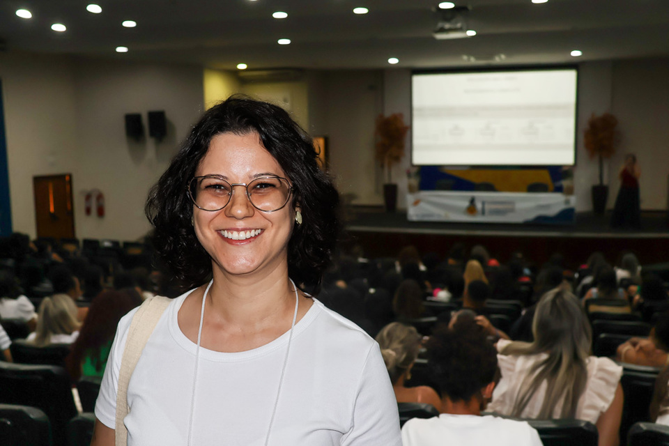 Mulher jovem, de pele branca e óculos sorri pra foto com auditório ao fundo